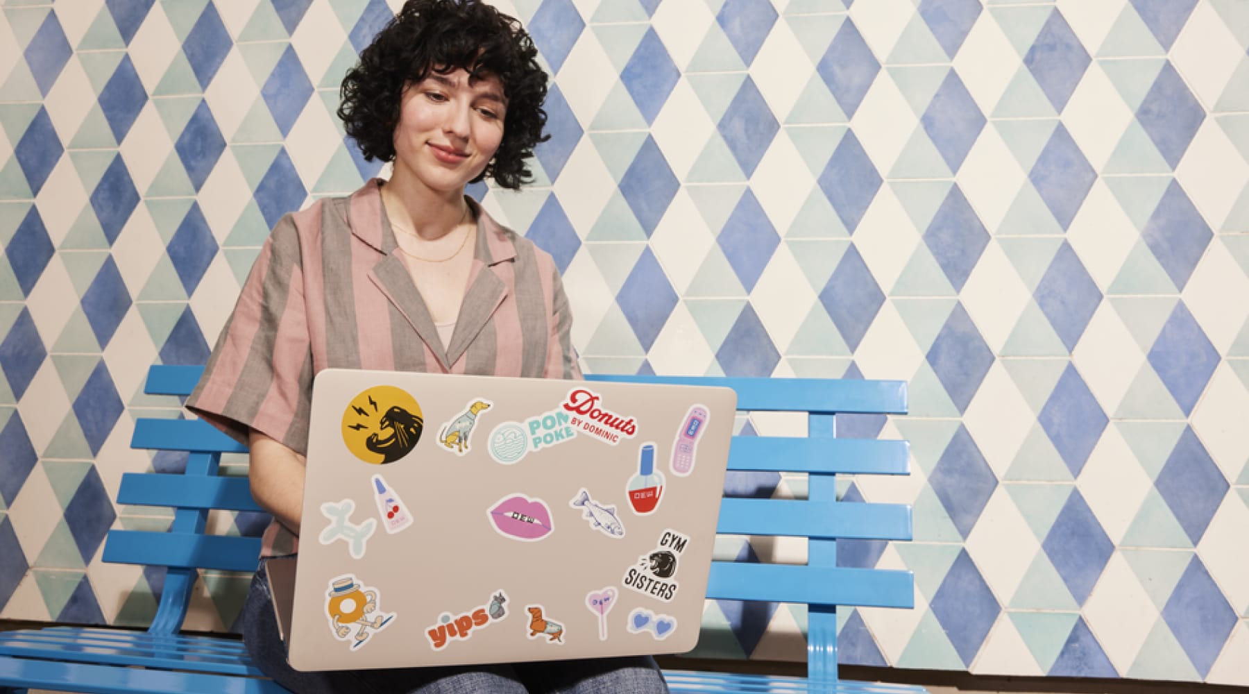 A person sitting on a bench on their laptop that's covered in custom stickers