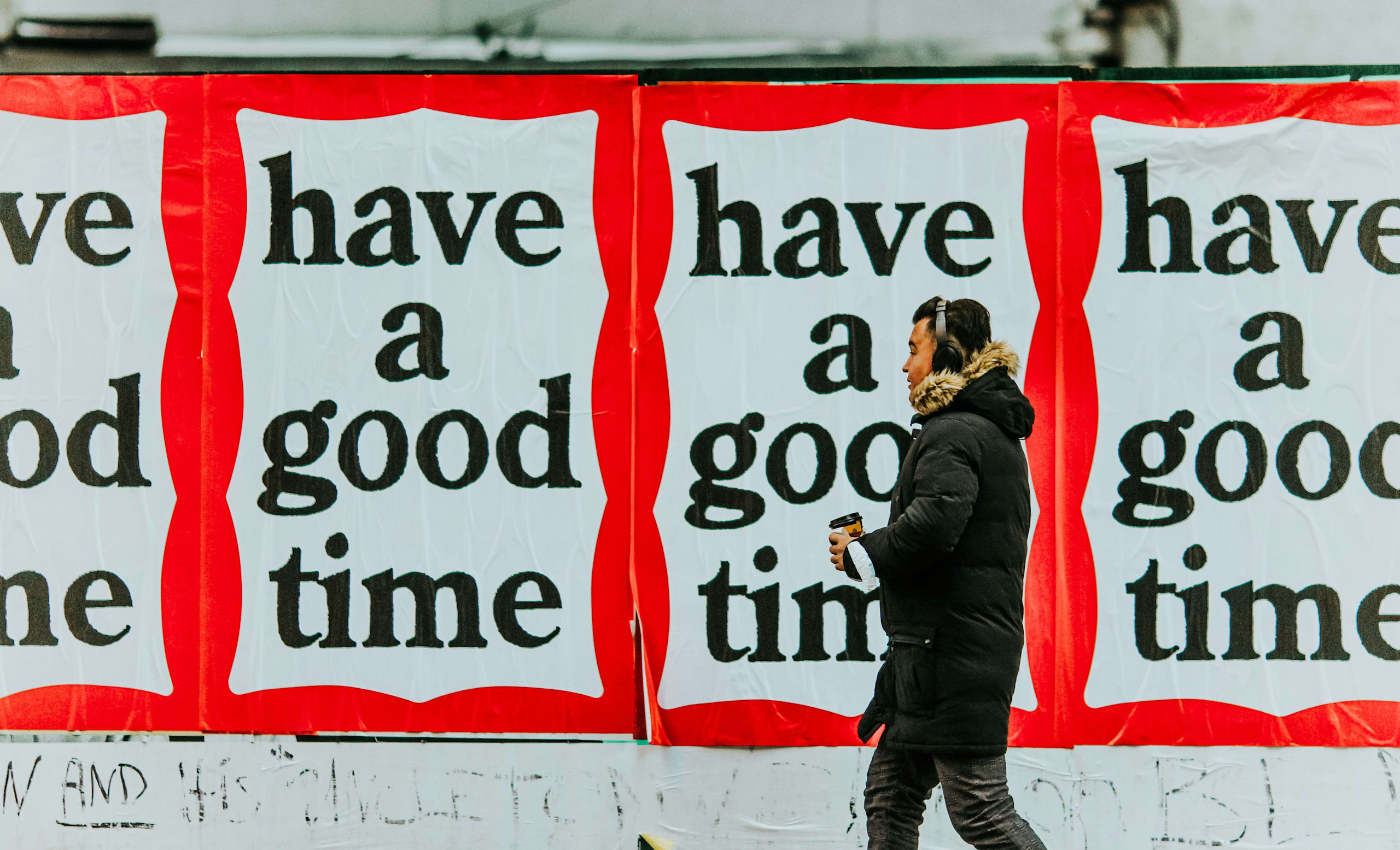 Person in black jacket holding coffee cup walking past a series of white posters with red borders with text “have a good time”