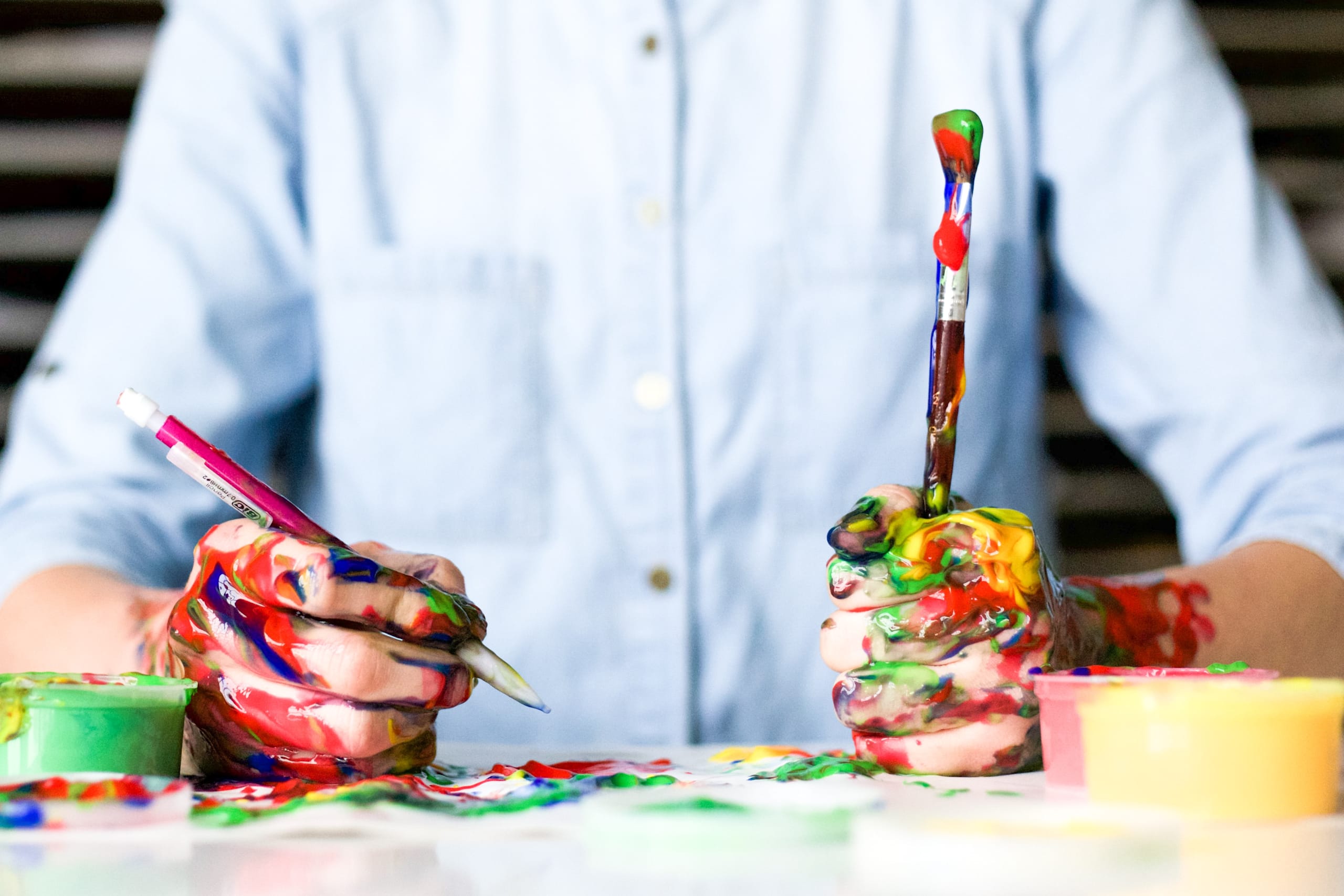 Nahaufnahme einer Person mit einem Stift und einem Pinsel in den mit Farbe bekleckerten Händen