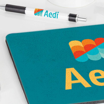 Branded merchandise on a white desk including a mug, USB stick, pen, and mousepad printed with a colorful logo