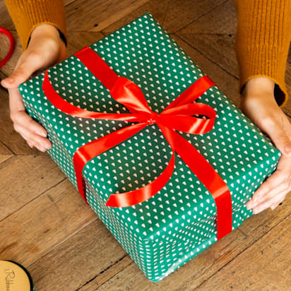 Two hands holding a green wrapped gift with a red bow on a wooden surface.