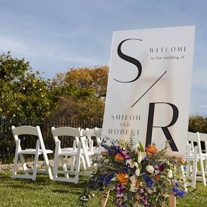 Wedding welcome sign with colorful floral arrangement on a wooden easel, set on a lawn with white ceremony chairs.
