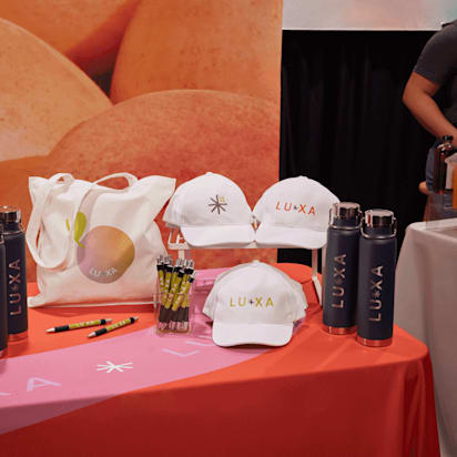 Colorful trade show table displaying branded swag items like hats, bottles, tote bags, and pens with a company logo.