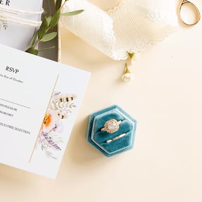 Elegant beige table with wedding RSVP cards on porcelain plate, decorated with roses, lace ribbons, and wedding rings.