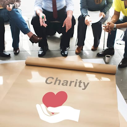 Multicultural group sitting in a circle around a large charity poster with a hand and heart logo.