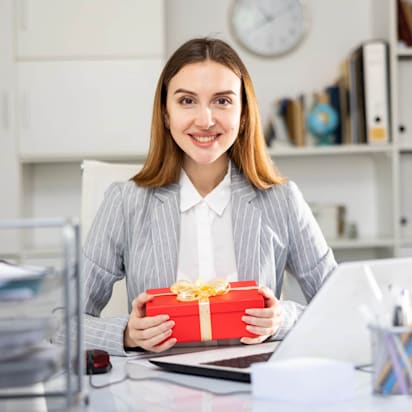 Elegant woman in an office holding a red gift box with a golden ribbon, smiling – the perfect employee appreciation gift.