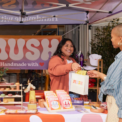 A vibrant craft fair stand where a smiling vendor hands a branded shopping bag to a happy customer. A real-world example of how to do market research for a small business.