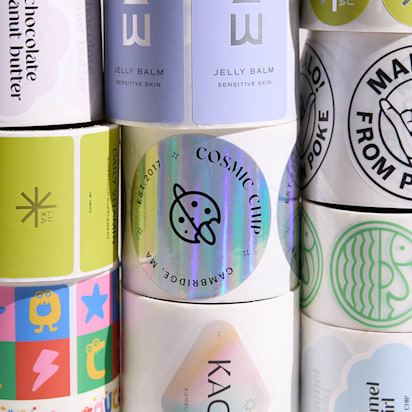 Multiple rolls of colorful custom stickers and logo labels stacked in a grid pattern, showing a variety of brand logos and designs.