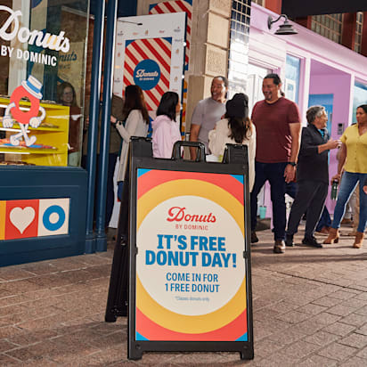 Colorful A-frame sign outside a donut shop with people lined up to enter.