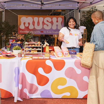 Outdoor fair stand showcasing signage in retail stores, with coordinated banner, vertical flag, A-sign and smiling vendor and customer.
