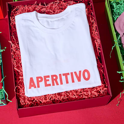 Three products in festive packaging, with the visible branding "Apertivo", are laid out side by side on a red table: A beanie, a thsirt and a cap