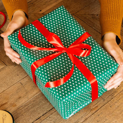 Zwei Hände halten ein grün verpacktes Geschenk mit roter Schleife auf einer Holzoberfläche.