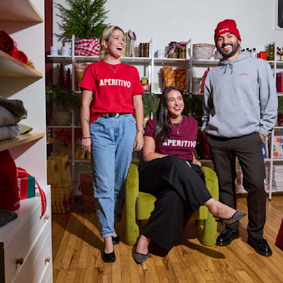 Business holiday prep example: three smiling people wearing branded Aperitivo accessories, t-shirts, and promotional materials in the background.