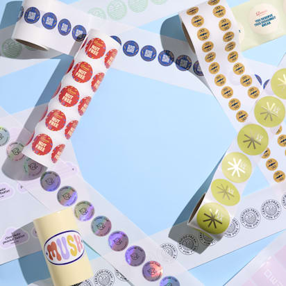 Various custom logo stickers on rolls in multiple styles, colors and finishes arranged on a pastel blue surface