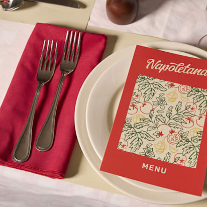 A restaurant table setting with a red menu titled “Napoletana” placed on a white plate, next to red napkin and silver cutlery.