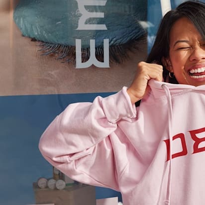Woman smiles as she pulls a pink branded hoodie down over her head