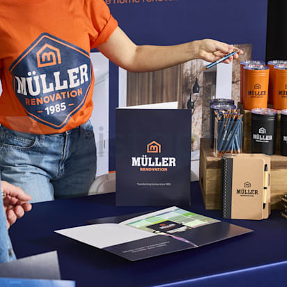 A branded collection of swag and merch from a Muller renovation company, notebook, pencils, drink containers, baseball hats and more arranged in layers across on a surface.