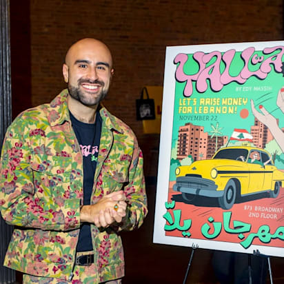 Man in colourful clothing stands smiling beside a large colourful poster advertising a community festival that he helps to organise - Yalla