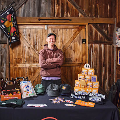 Direct photograph of the featured artist/designer, Kirk Wallace, standing behind a table featuring multiple example of his work with VistaPrint products eg. apparel, mugs, packaging, stickers