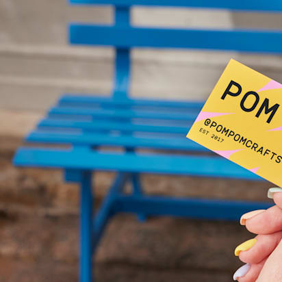 Hand holding a colorful POM POM business card in front of a bright blue bench.