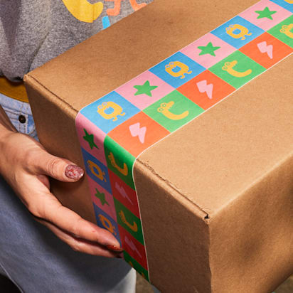 Brown cardboard shipping box sealed with colorful custom packaging tape featuring playful graphics, held by a person.