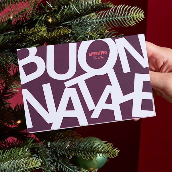 Hand holding a typographic Christmas card reading “Buon Natale” in front of a Christmas tree.