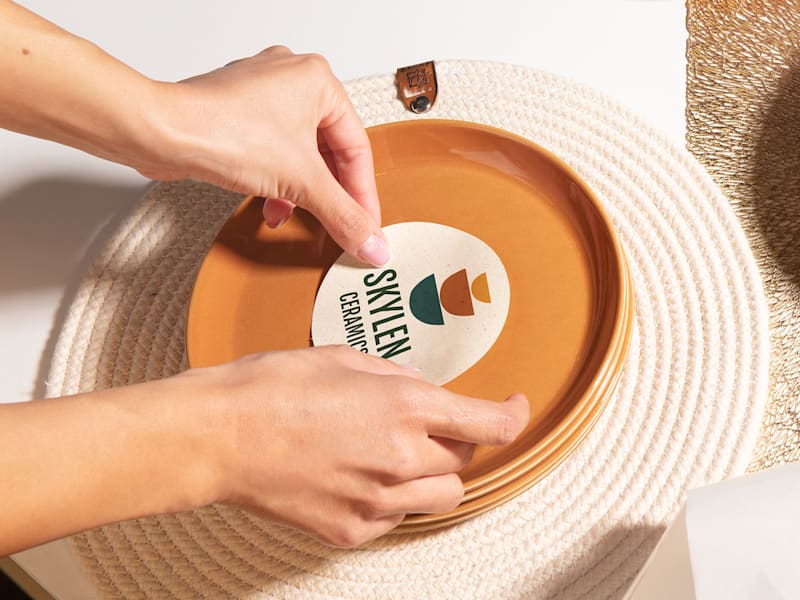 Hands applying a ceramic brand sticker to a red plate, showcasing logo design software in action.