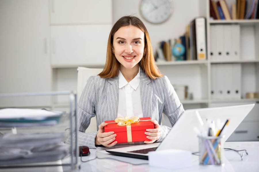Elegant woman in an office holding a red gift box with a golden ribbon, smiling – the perfect employee appreciation gift.