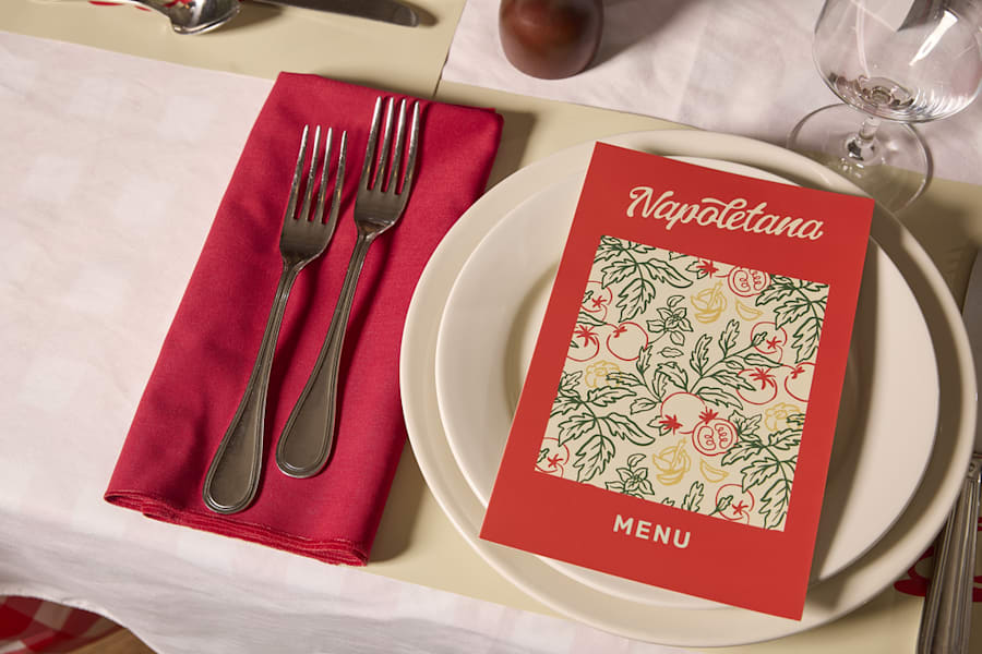 A restaurant table setting with a red menu titled “Napoletana” placed on a white plate, next to red napkin and silver cutlery.