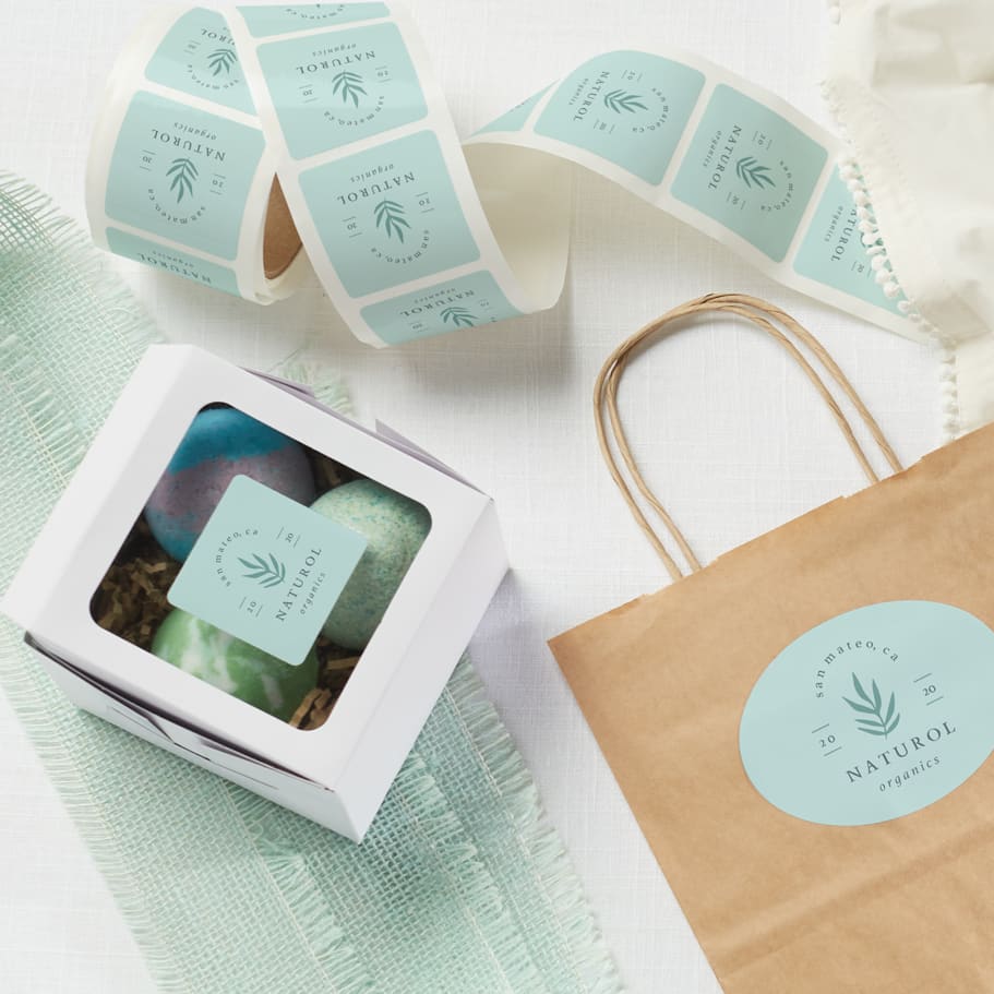 A roll of a small business’s custom stickers in light blue, laid across a table with product boxes and kraft paper bags around