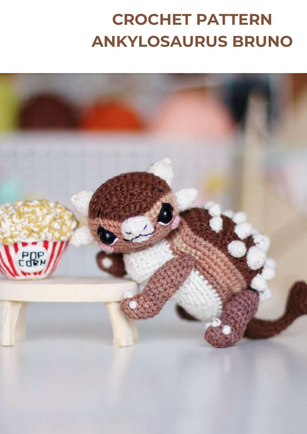 Brown and cream crochet ankylosaurus with white spikes next to a crochet popcorn bucket