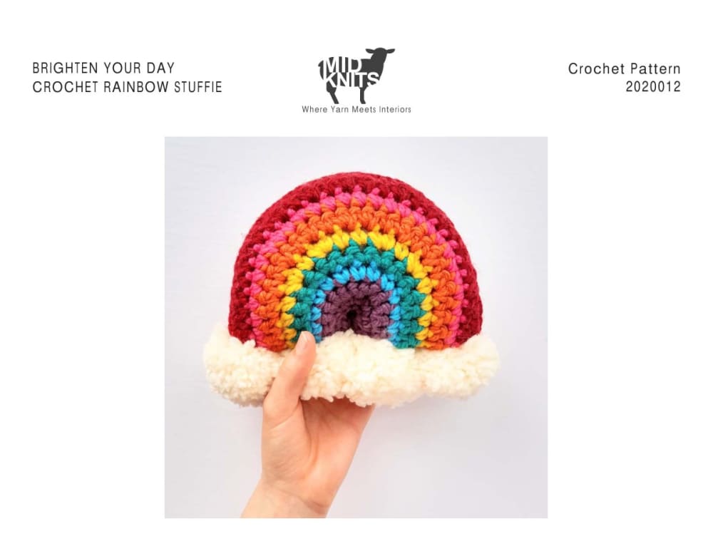 Hand holding a bright crochet rainbow stuffie with white pom-pom clouds at both ends