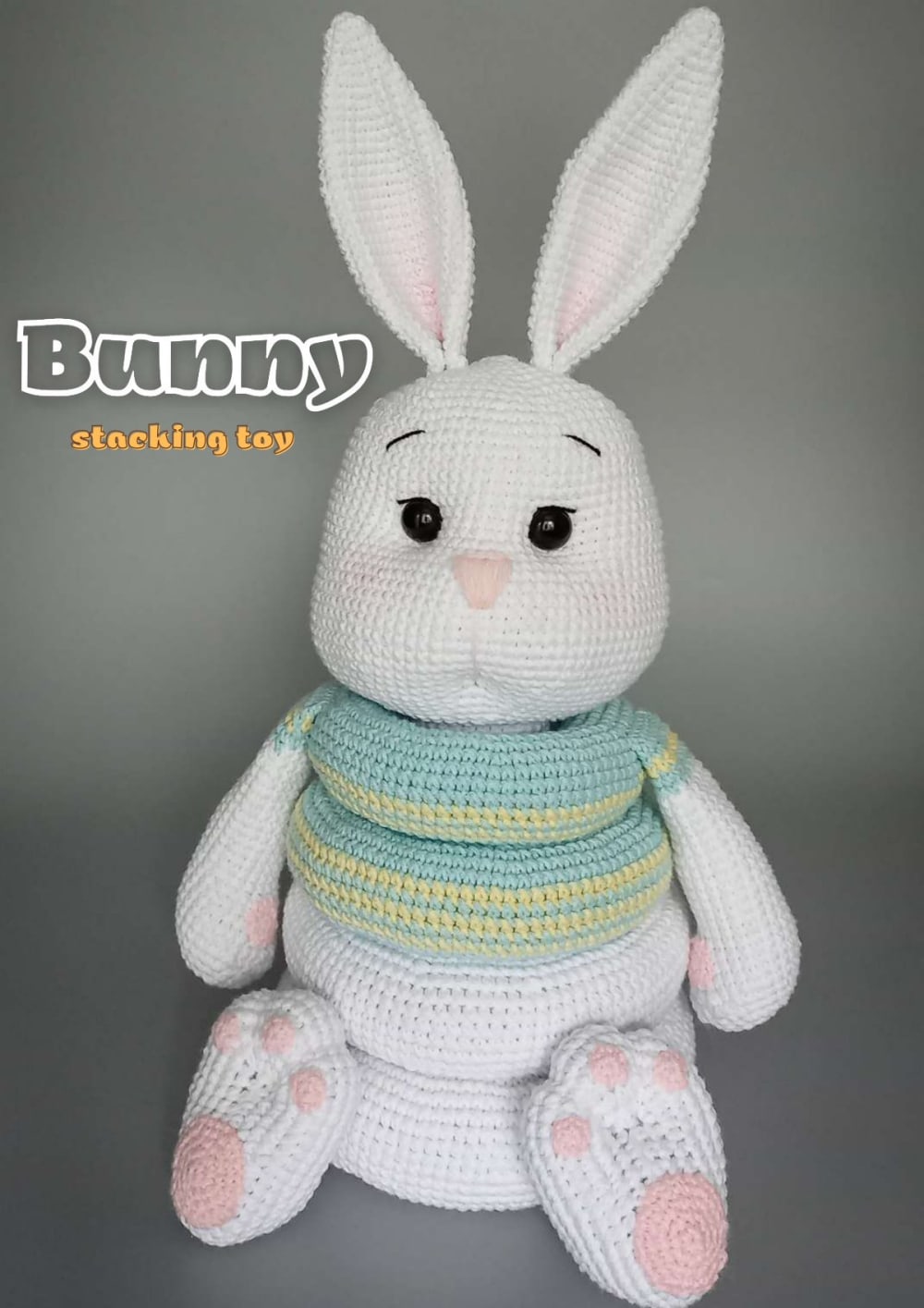 White crochet bunny stacking toy with pink ears and paws and a striped turquoise and yellow ring on its body