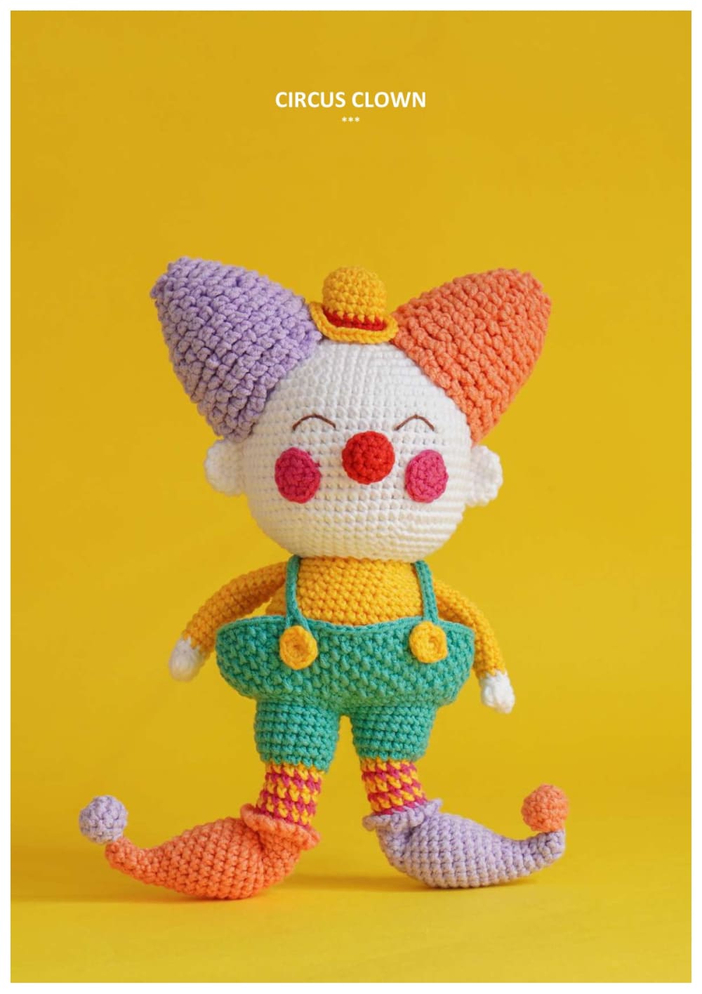 Amigurumi circus clown with a purple and orange pointed hat, red nose, yellow shirt and green overalls on a yellow background