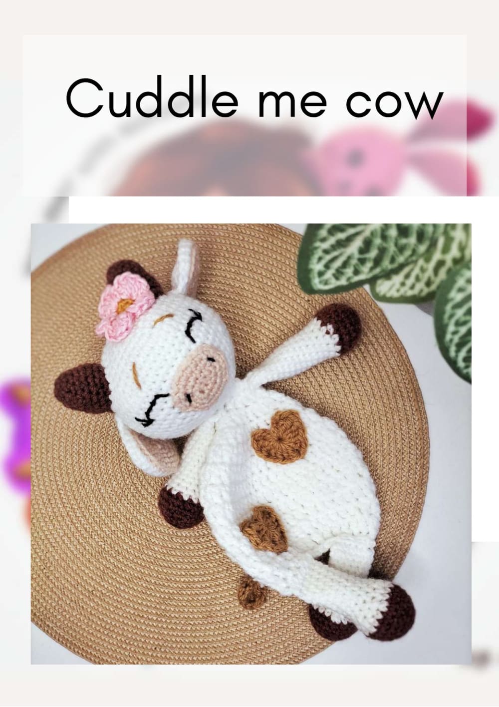 White crochet cow lovey with brown heart spots, brown hooves and a red flower lying on a round woven mat