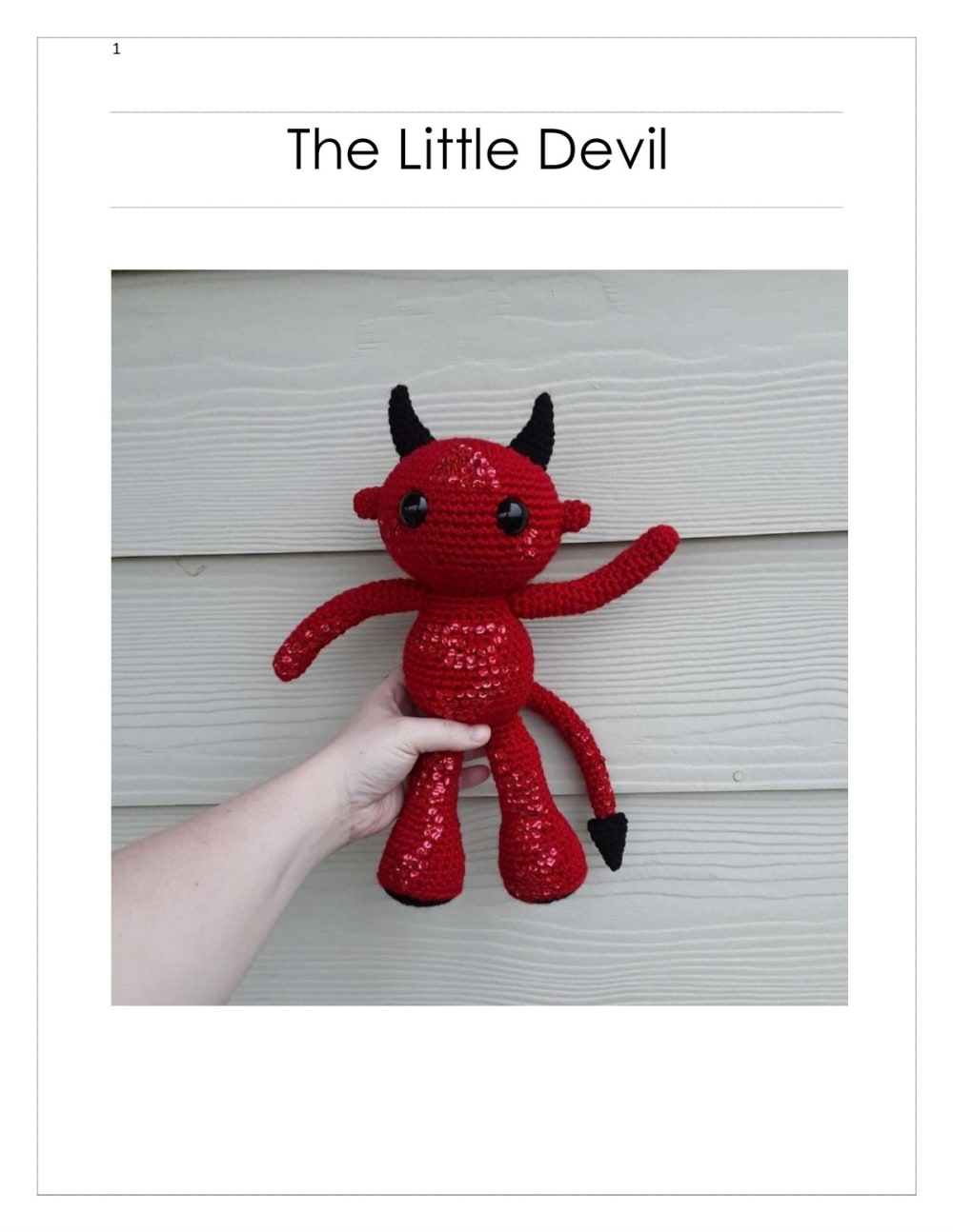 Red crochet little devil with black horns, black eyes, thin arms and a pointed black tail held in a hand