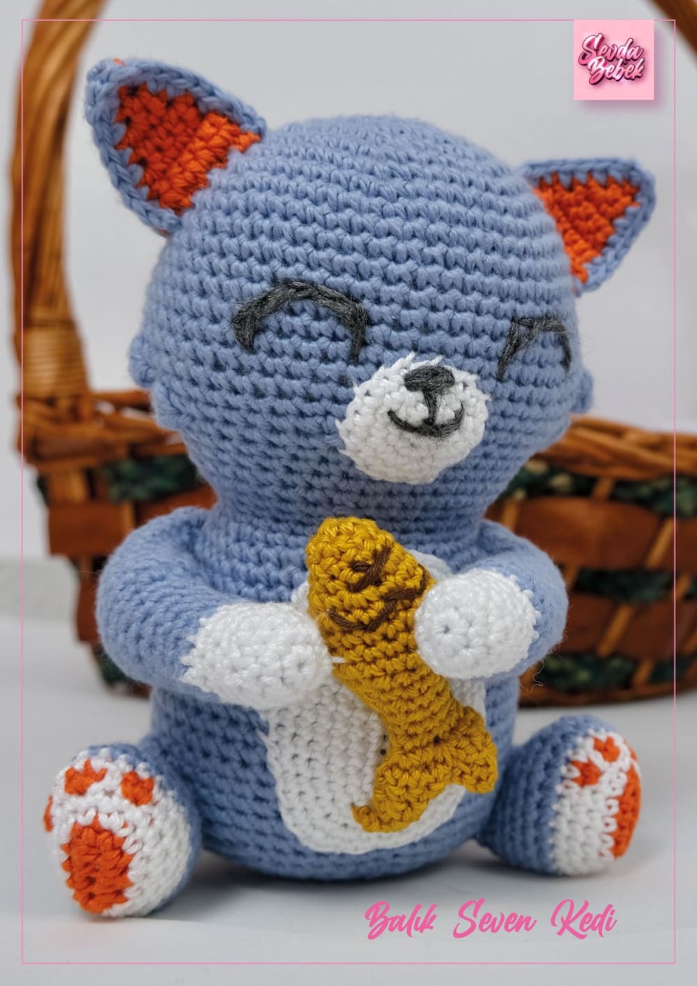Sleepy blue crochet cat sitting in a basket holding a yellow fish