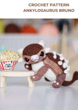 Brown and cream crochet ankylosaurus with white spikes next to a crochet popcorn bucket