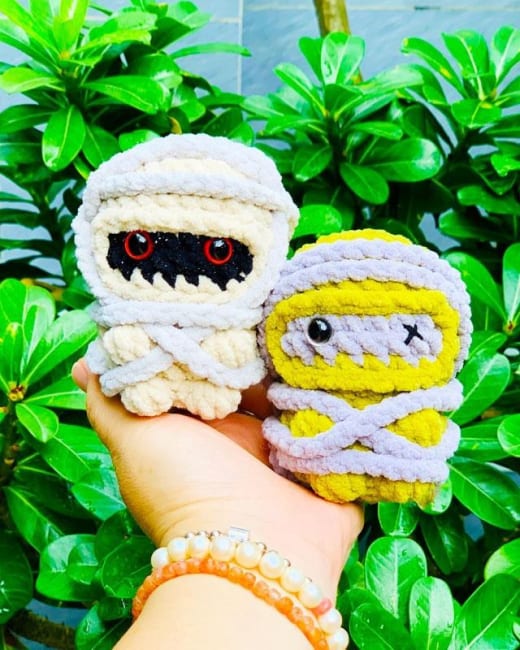 Crochet mummy pattern: two finished amigurumi mummy dolls, one white and one yellow, held in a hand against green leaves