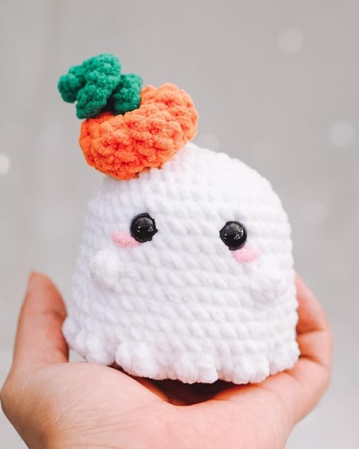 Crochet ghost pattern: white amigurumi ghost wearing a small orange pumpkin hat with a green leaf, held in a hand