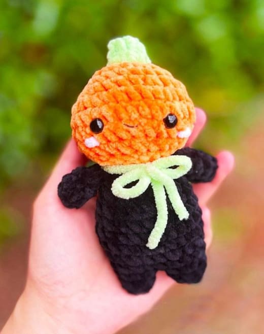 Pumpkin amigurumi doll pattern: crochet doll with an orange pumpkin head, black outfit and green bow held in a hand outdoors