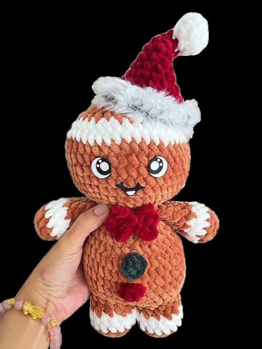 Gingerbread santa amigurumi: crochet gingerbread man in a red and white Santa hat and red bow tie held in a hand against a black background