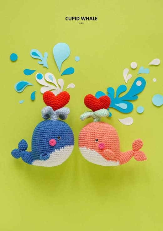 Two crochet cupid whales in blue and coral with red hearts on a yellow background
