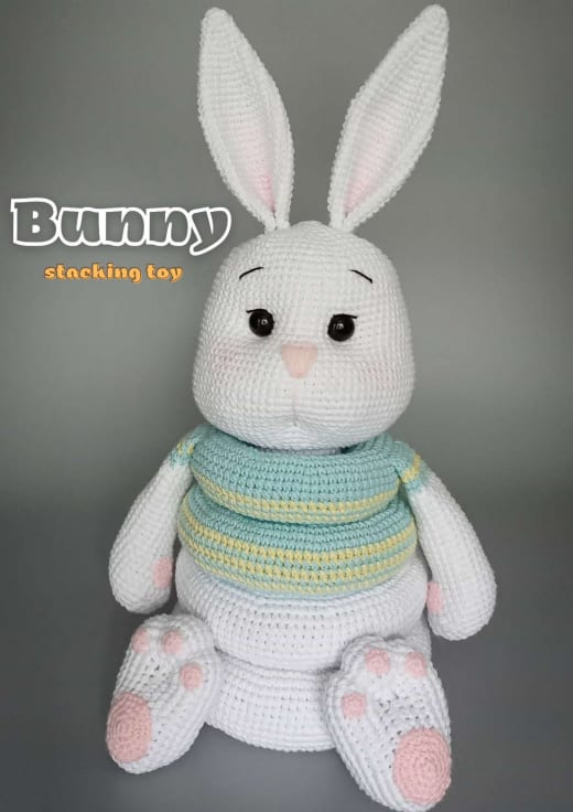 White crochet bunny stacking toy with pink ears and paws and a striped turquoise and yellow ring on its body