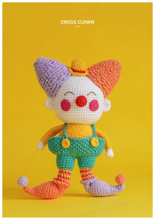 Amigurumi circus clown with a purple and orange pointed hat, red nose, yellow shirt and green overalls on a yellow background