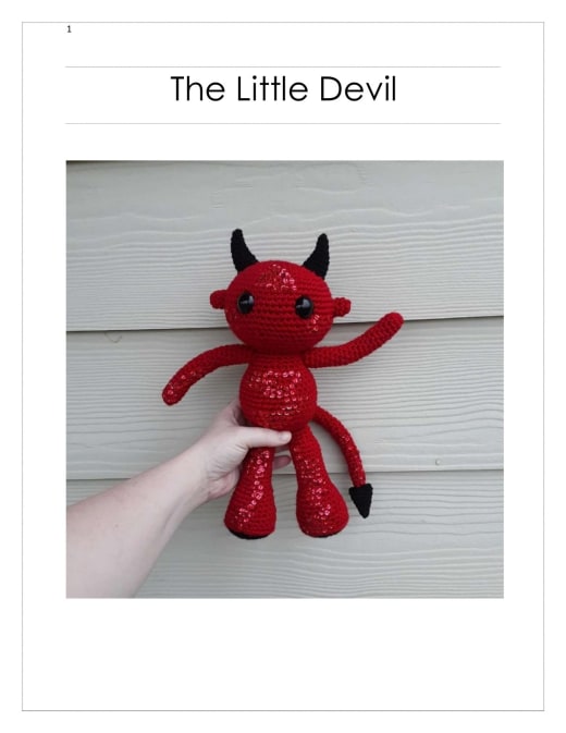 Red crochet little devil with black horns, black eyes, thin arms and a pointed black tail held in a hand