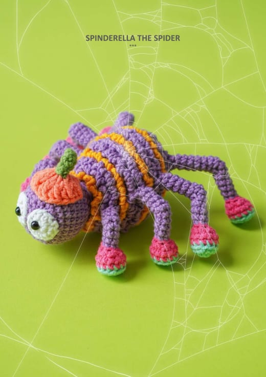 Purple and orange crochet spider with big white eyes, thin legs and pink feet on a green background