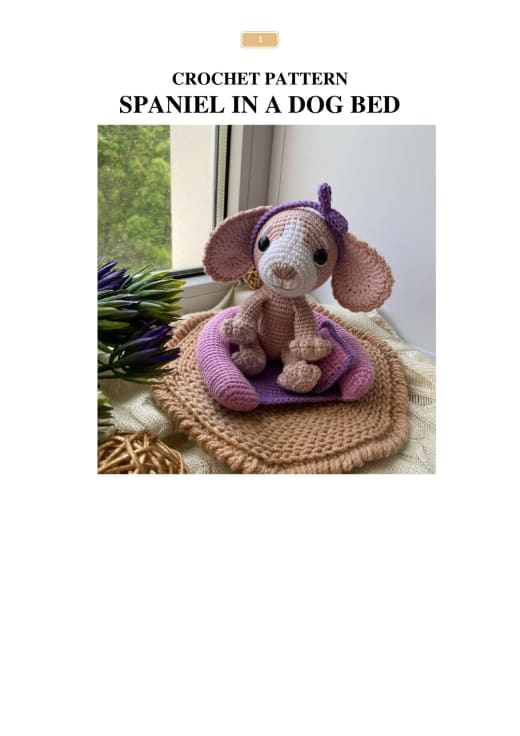 Pink and beige crochet spaniel with floppy ears and a purple headband sitting on a purple dog bed with a pillow