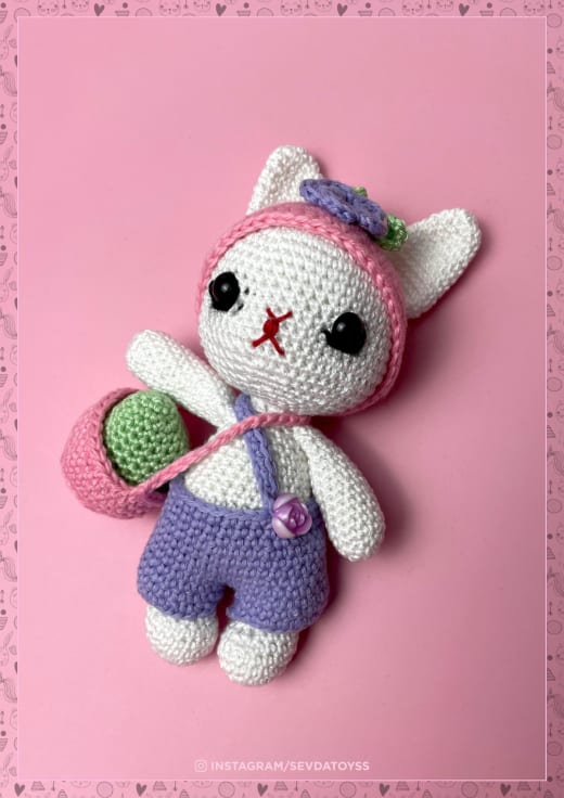 White crochet kitty in a pink bunny-ear hat and purple shorts holding a green egg