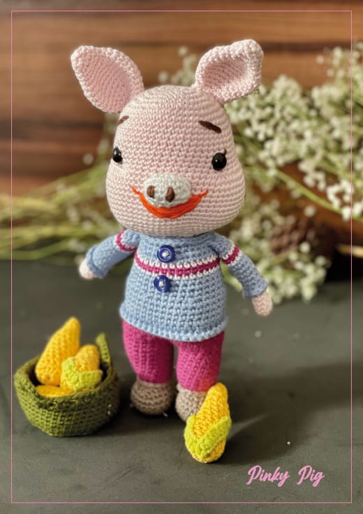 Pink crochet pig in a striped top and pants standing beside a basket of yellow eggs